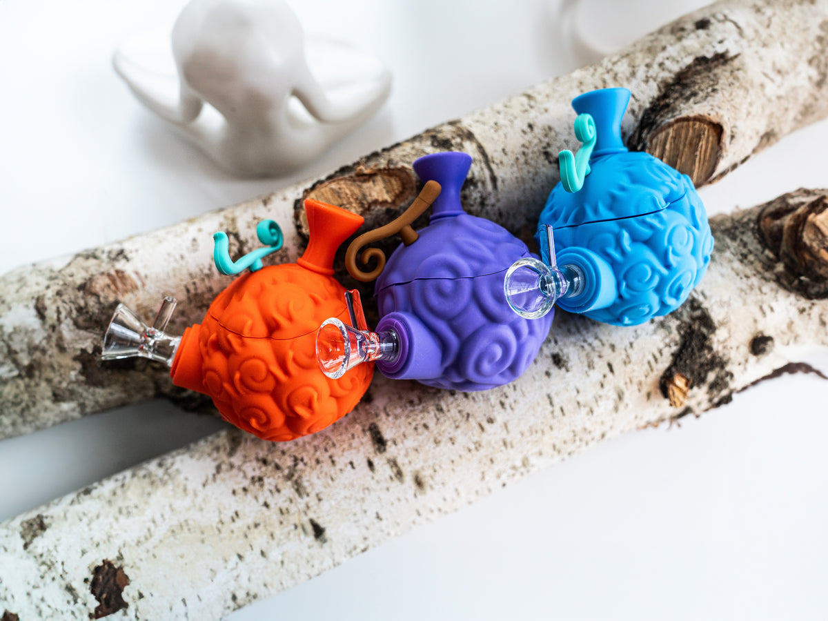 Orange, purple, and blue devil fruit silicone water pipes with removable glass bowls on birch logs