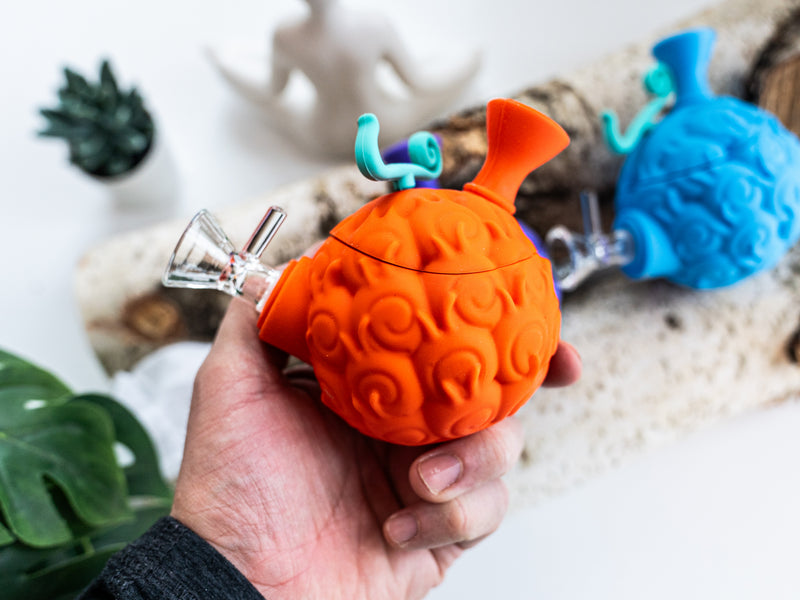 Orange devil fruit silicone water pipe held in one hand with a blue pipe in the background
